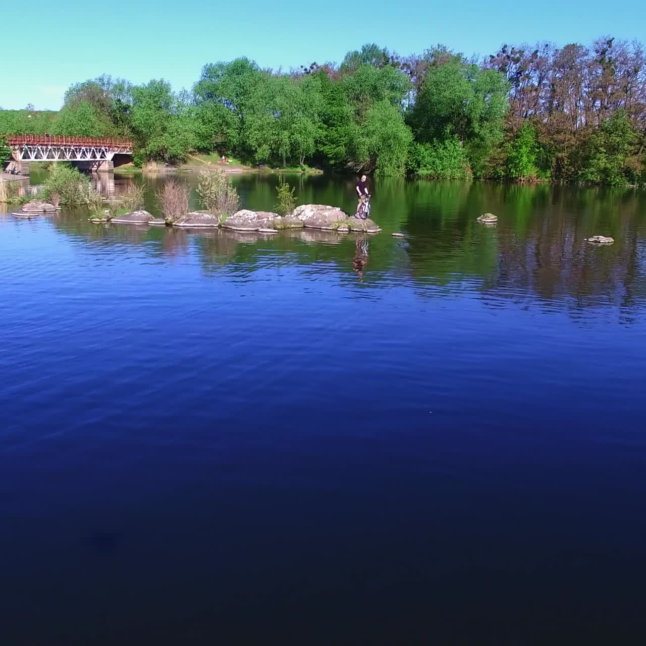 Beautiful natural view on bright sunny summer day. Drone flying over the calm blue river with green wooded waterfront. People spending time in the nature outside the city