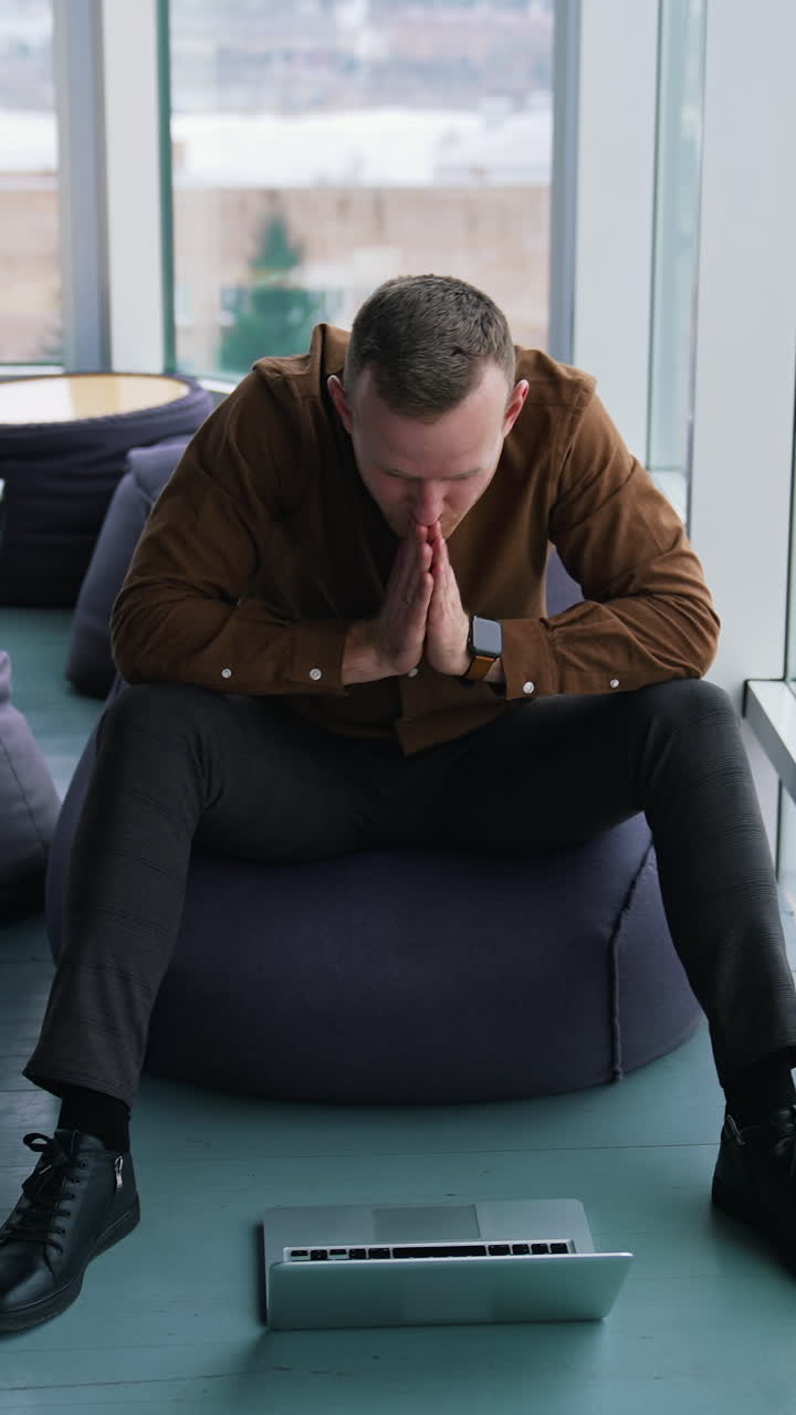 Young man with a laptop near the window. Handsome guy in shirt is sitting on a comfortable ottoman and looking on his laptop waiting for something. Vertical video