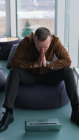 young man with a laptop near the window. handsome guy in shirt is sitting on a comfortable ottoman and looking on his laptop waiting for something. vertical video
