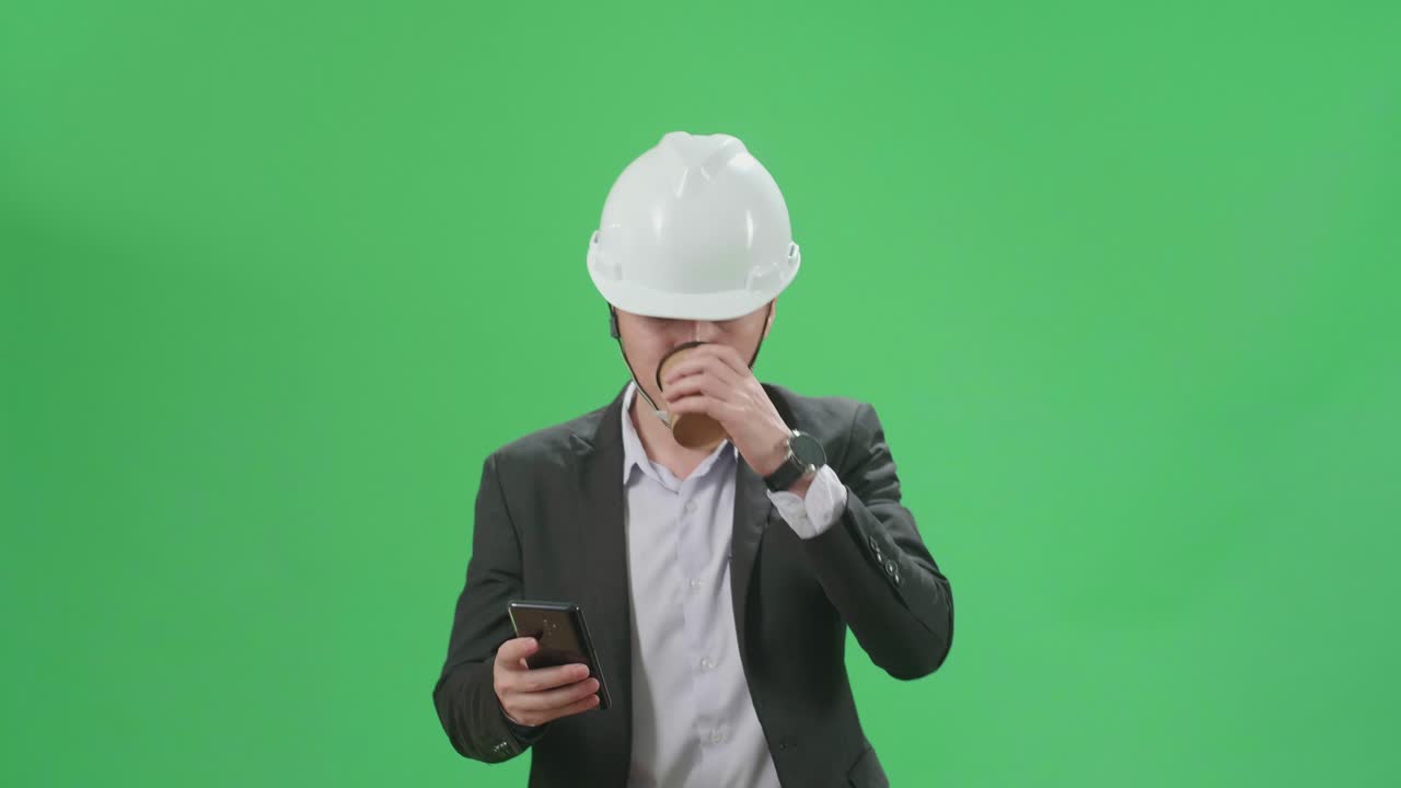 Asian Chief Engineer In The Hard Hat Use Mobile Phone And Drink Coffee While Walking On Green Screen Chroma Key