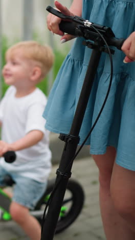 una mujer monta un scooter en un puente, mirando pensativo mientras un niño joven la sigue en una bicicleta