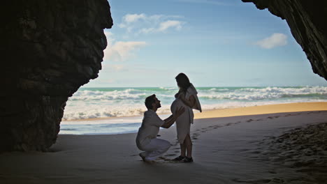 expectant father stroking pregnant tummy wife standing in beach rock shadow.
