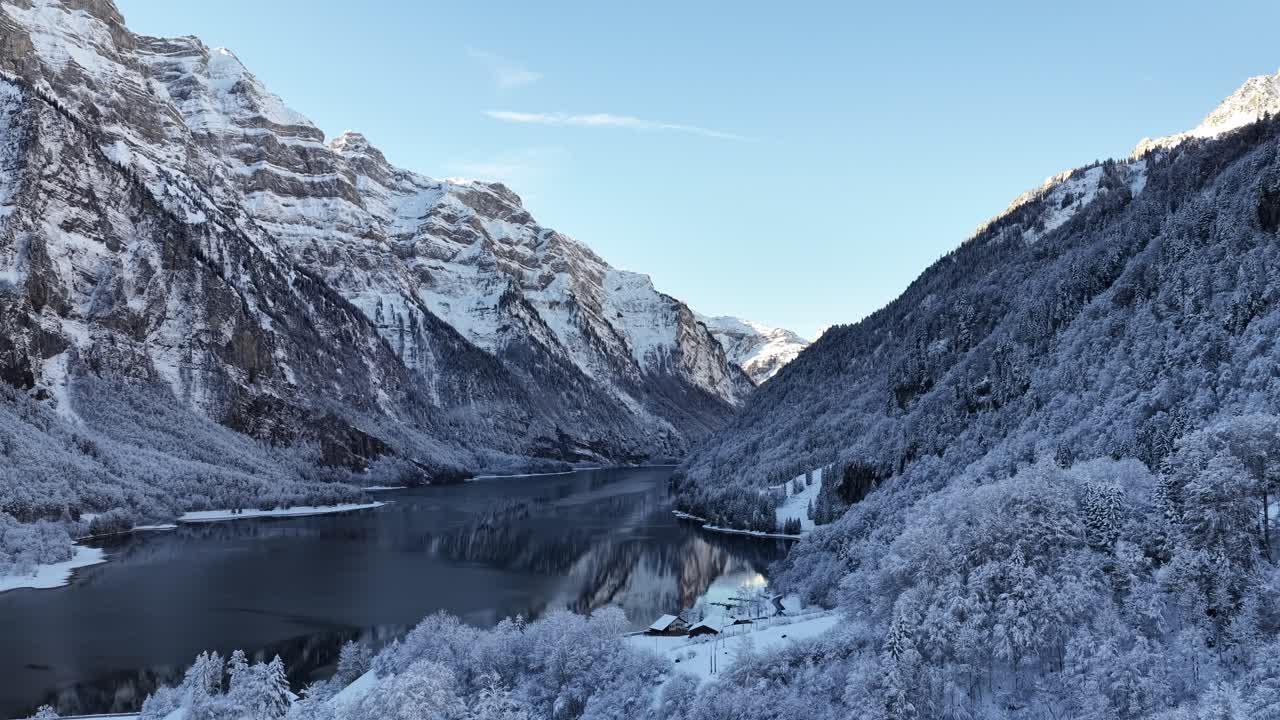 Ilmakuva Klöntalersee-järvestä, Glarus, Sveitsi. Lumipeitteiset vuoret ja kuuraiset puut reunustavat rantoja heijastuen tummasta, tyynestä järvivedestä. Kylmä, majesteettinen alppimaisema.