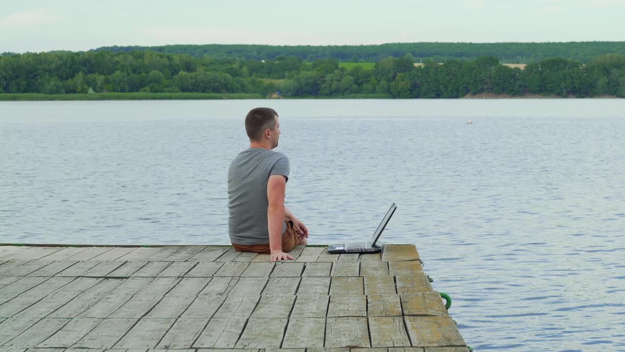Handsome man sitting at the edge of the bridge finishes typing on a laptop and enjoys the natural background. Work with pleasure outdoors near the river
