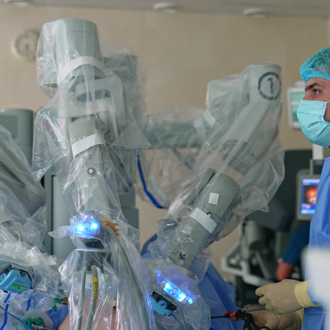 Young male doctor stands near the patient operated by Da Vinci robot. Specialist watching the surgery process at the screen holding a medical tool