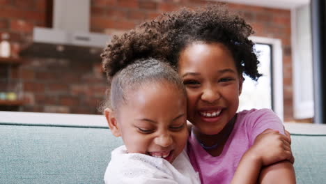 Portrait-Of-Two-Sisters-Sitting-On-Sofa-At-Home-Smiling-Into-Camera