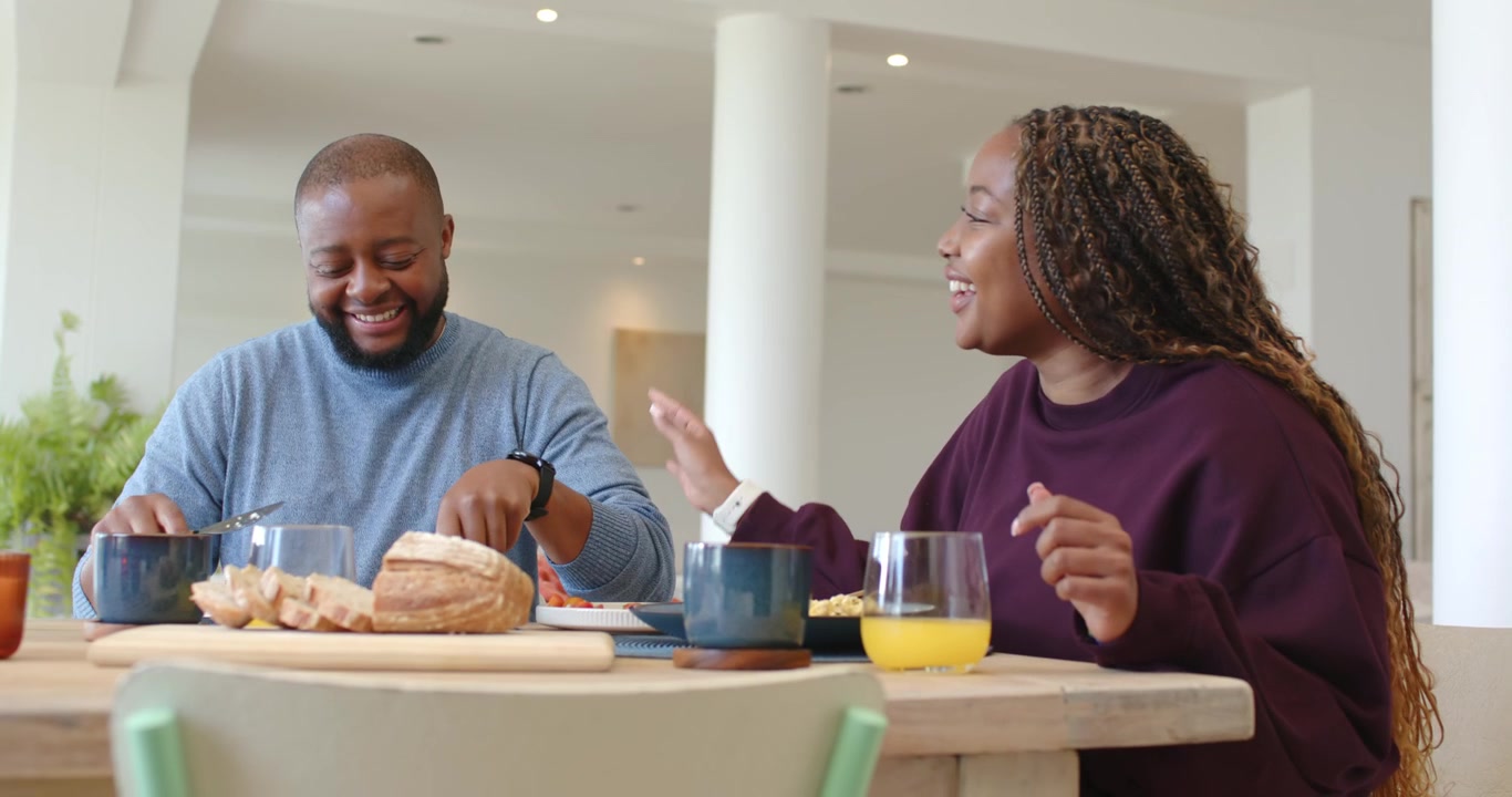 African American couple sitting at table, woman talking causing man slicing bread for breakfast
