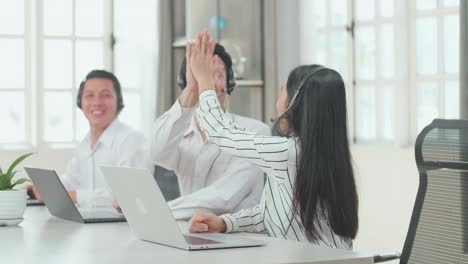 Three Asian Call Center Agents Wearing Headsets Sitting On The Chairs And Being Happy Due To Success Working With Computers At The Office