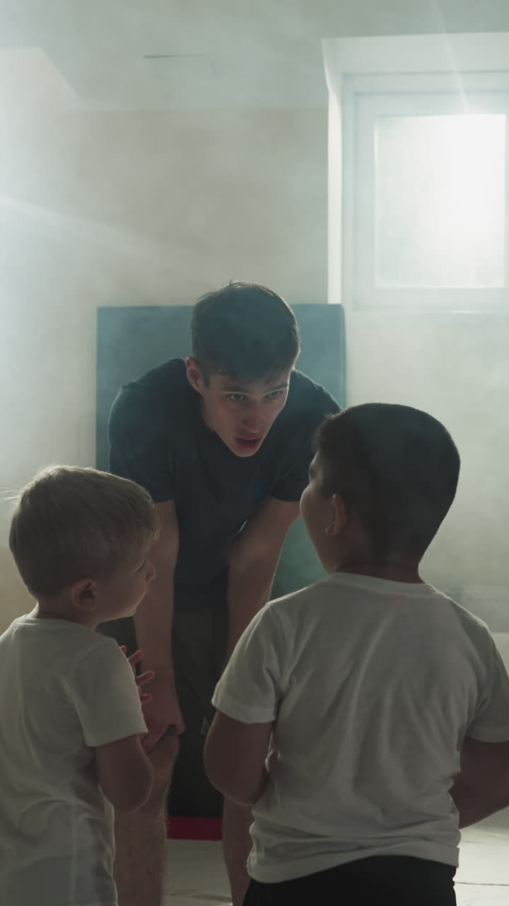 어린이 그룹이 무술 수업에서 센세이를 듣고 있습니다. 작은 전투 학생들은 체육관에서 느린 동작으로 운동을 준비하기 위해 강사 앞에 손을 흔들고 있습니다.