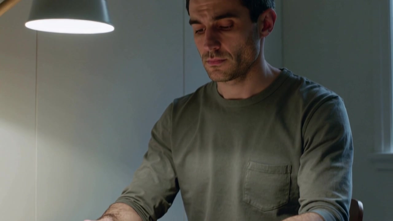 Câmera recuando, revelando um homem de camisa verde-oliva lendo, virando um livro em uma escrivaninha de estudo sob a lâmpada.