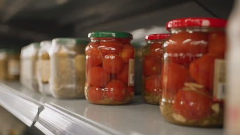 close up of young mother hands taking transparent glass jar of tomatoes from store shelf rails in bright grocery aisle highlighting manicure polished nails wrist watch and blurred retail background