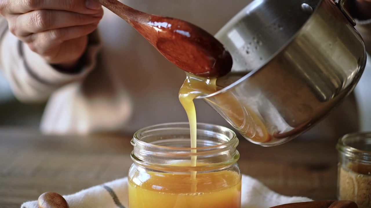Delle mani inclinano un pentolino, versando del liquido in un barattolo largo sul bancone della cucina, con un cucchiaio che guida il flusso per la conservazione.