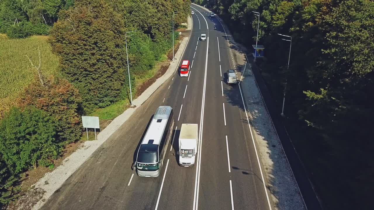 New road with four lines and the cars going by in summer. Green trees growing on roadside of a two-lane road. Bus stop from the right side of the road. Aerial view along the highway