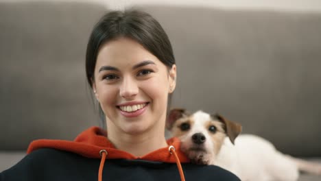 brunette woman sitting on floor near sofa, small dog jack russell terrier on her shoulder looking to the camera