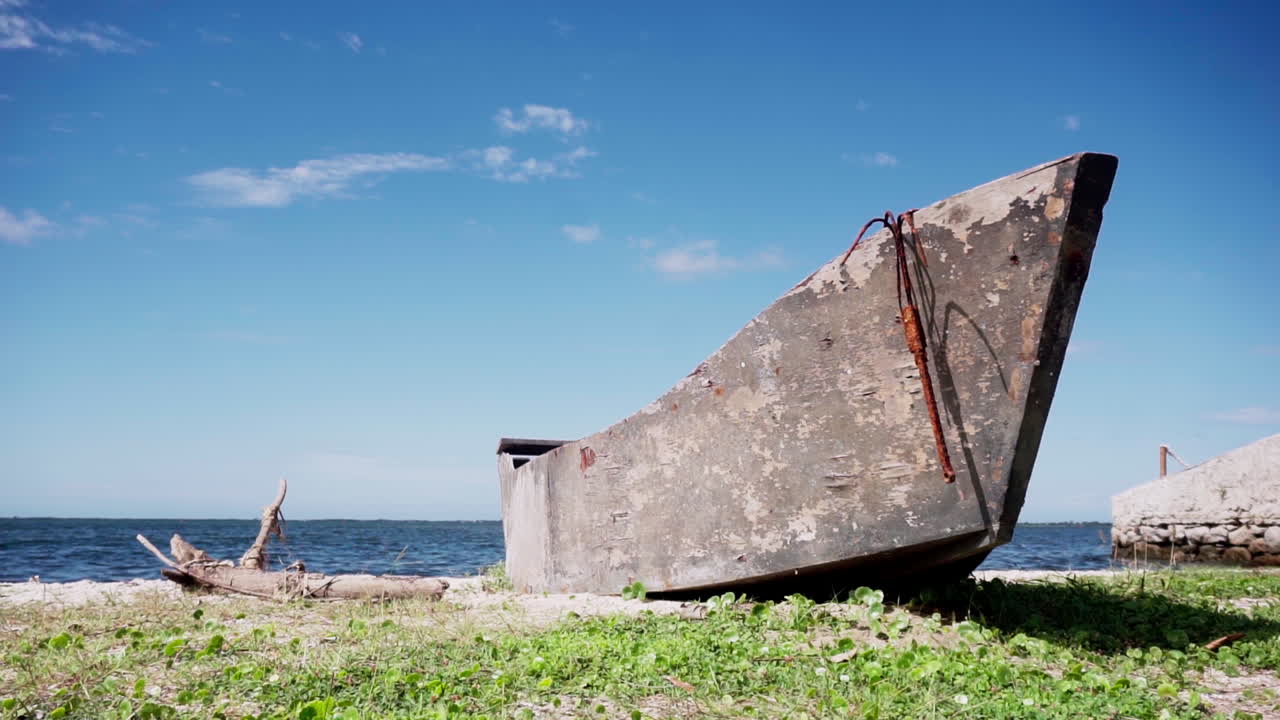 A rusted boat with a rusted anchor lies on the shore of Araruama, Brazil, surrounded by green grass and calm blue waters, under a clear sky, evoking a sense of abandonment.