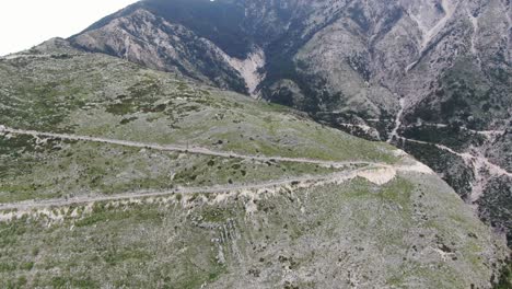 Drone-view-in-Albania-flying-over-a-green-and-rocky-mountain-with-serpent-road-climbing-a-port-on-a-cloudy-day