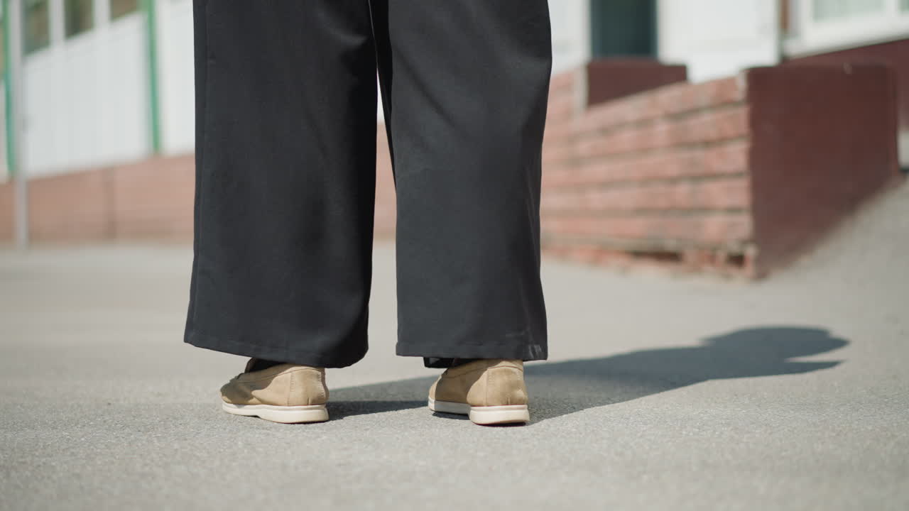 Person stands quietly on bright pavement wearing black wide pants and brown shoes, shifting slightly under sunlight, subtle movement and shadows create calm mood of waiting