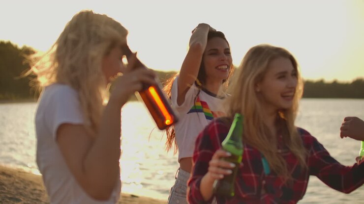 Girls carefree and cheerful move young bodies on the beach with beer on the open air party near the sand river coast in summer evening.