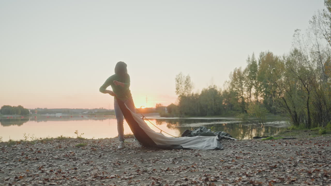 Weitwinkelaufnahme einer weißen Frau, die bei Sonnenuntergang am See ein Zelt aufbaut. Ganzkörpersilhouette am blätterbedeckten Ufer, beim Spannen der Zeltleinwand und Befestigen der Heringe. Weiches goldenes Licht auf ruhigem, spiegelndem Wasser. Eine alleinreisende Camperin bereitet den Abend vor.