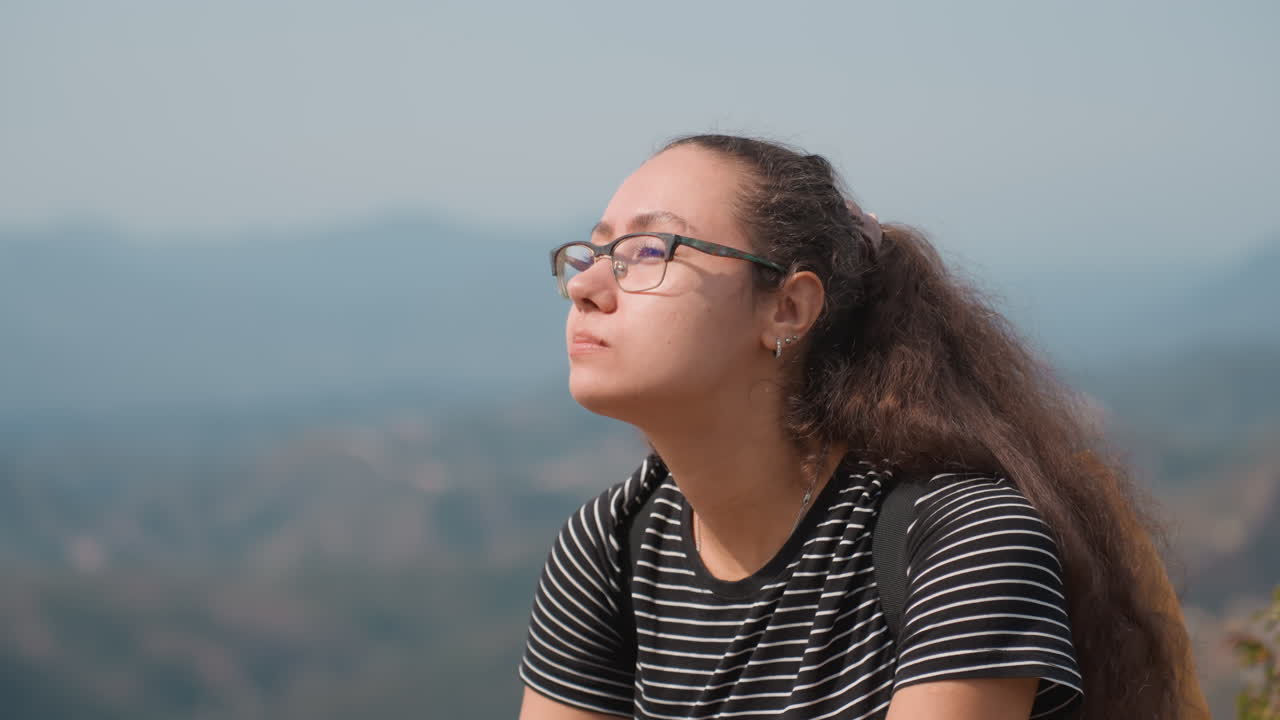 Young woman tasting apple with mountain view, closeup portrait, glasses and ponytail, striped tee, distant ridgelines and historic tower, soft breeze, thoughtful expression, quiet travel reflection