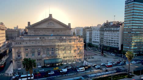 aerial panning view capturing colon theater on the rear entrance, busy downtown rush hour traffics on 9 de julio avenue with big glowing sun in the background at sunset, buenos aires city, argentina