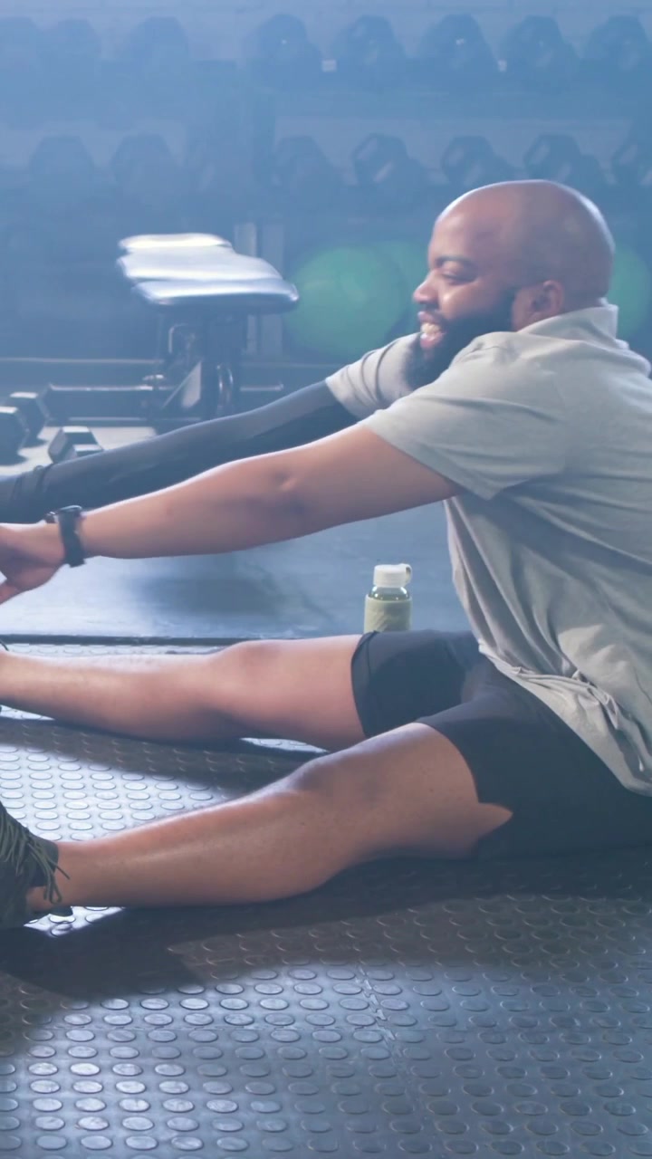 Video verticale: Uomo calvo in abbigliamento sportivo che si allunga toccando le punte dei piedi su un tappetino da palestra, con manubri e panca.