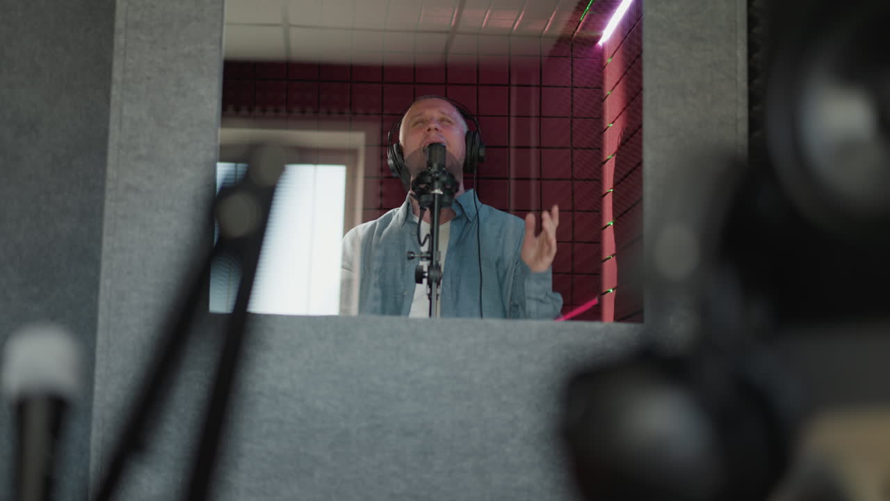 un hombre canta apasionadamente en un estudio de grabación, con auriculares negros y una camisa azul de manga larga. la escena captura su intensa expresión mientras canta con la boca abierta
