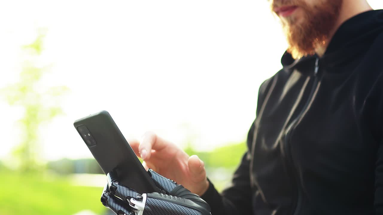 joven barbudo con una prótesis de brazo de metal al aire libre mirando la pantalla de su teléfono inteligente.