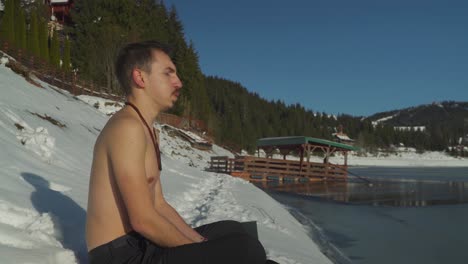 young man with no shirt sitting in snow beside frozen mountain lake breathing heavily