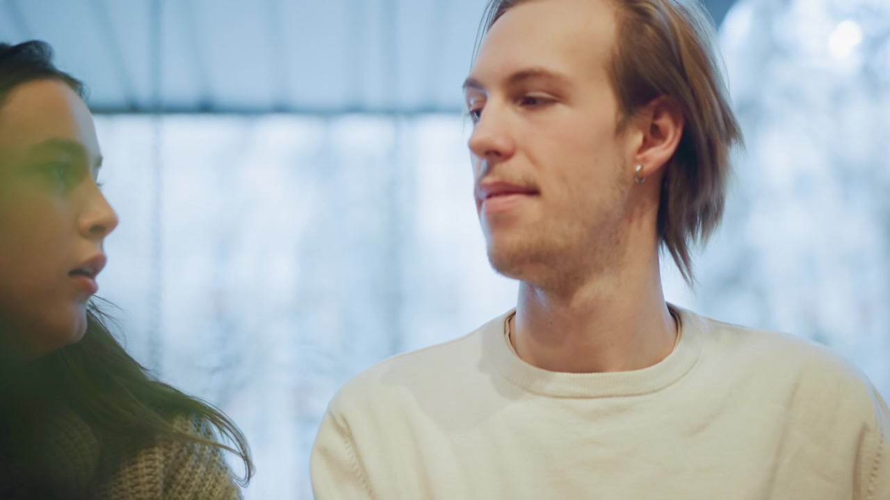 Nærbilde av kvinne og mann som sitter side om side i mykt lys, begge ser bort fra kameraet. Kvinnen har på seg en strikket genser, bakgrunnen har uskarpt grønt og lyseblå toner