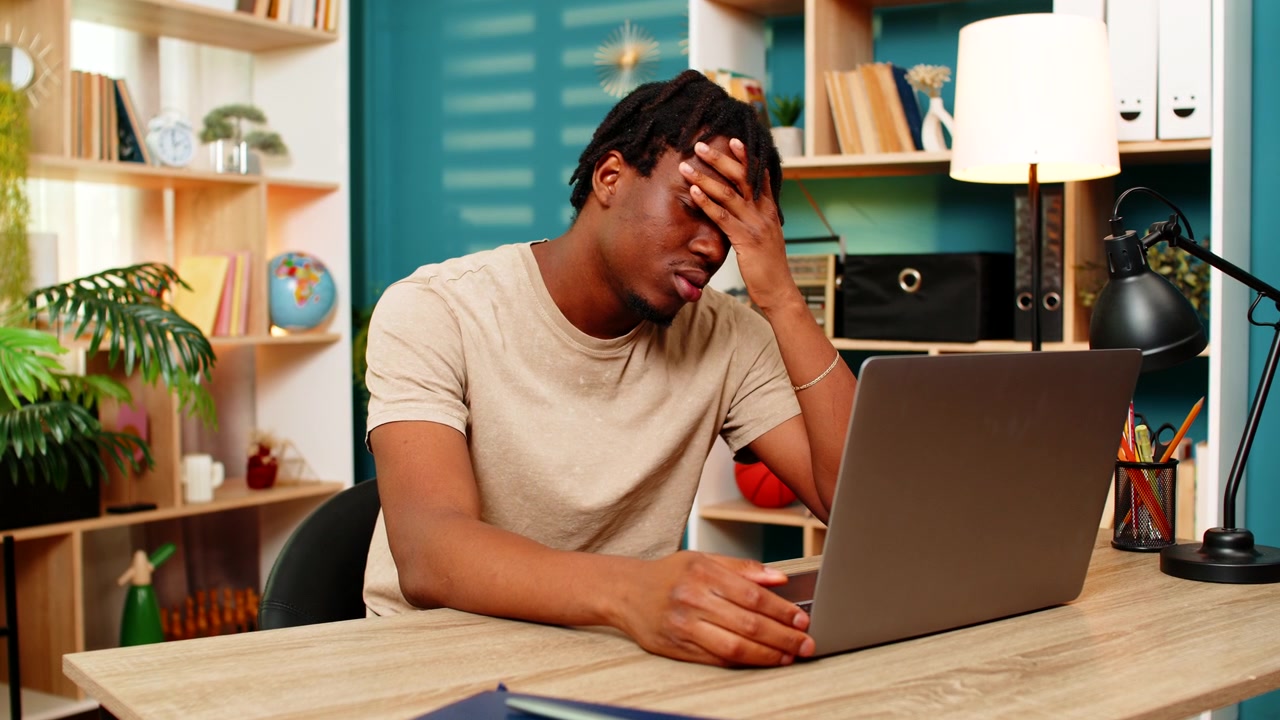 Afroamerikansk ung mann ved hjemmebordet, frustrert over en laptop-feil, føler stress etter avslag