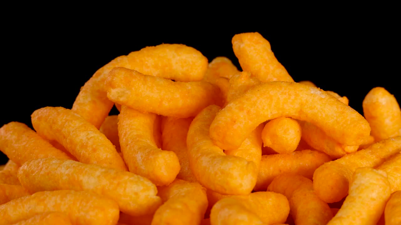 A camera smoothly zooms in on a pile of cheese puffs, brightly lit against a solid black background, highlighting texture and vibrant orange color