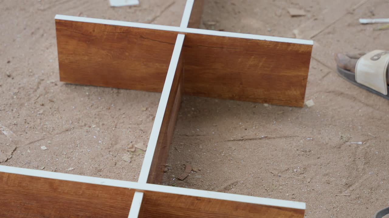 Disposizione a croce di legno sulla sabbia che mostra due assi bordate in melaminico che si intersecano su terreno polveroso, primo piano di un divisorio in truciolato con sottile segatura, giunzione precisa e posizionamento misurato per uno scaffale