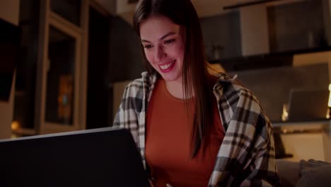 una chica morena alegre y segura en una camisa a cuadros y una camiseta naranja se sienta en un sofá gris y ve videos divertidos usando una computadora portátil en un apartamento de estudio moderno por la noche