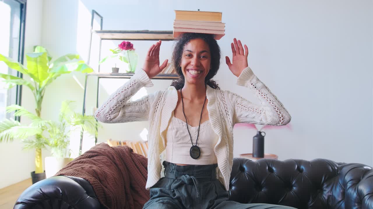joven feliz mujer afroamericana con libros en la cabeza se sienta en el sofá