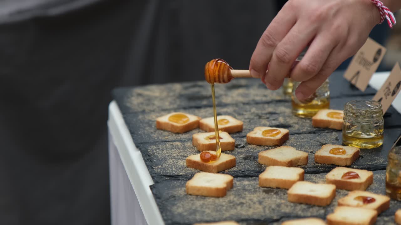 A hand is pouring golden honey through a dipper on taster crackers for people to try at local market stall.