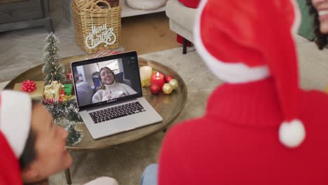 Familia-Diversa-Con-Sombreros-De-Santa-Usando-Una-Computadora-Portátil-Para-Una-Videollamada-Navideña-Con-Una-Mujer-Feliz-En-La-Pantalla