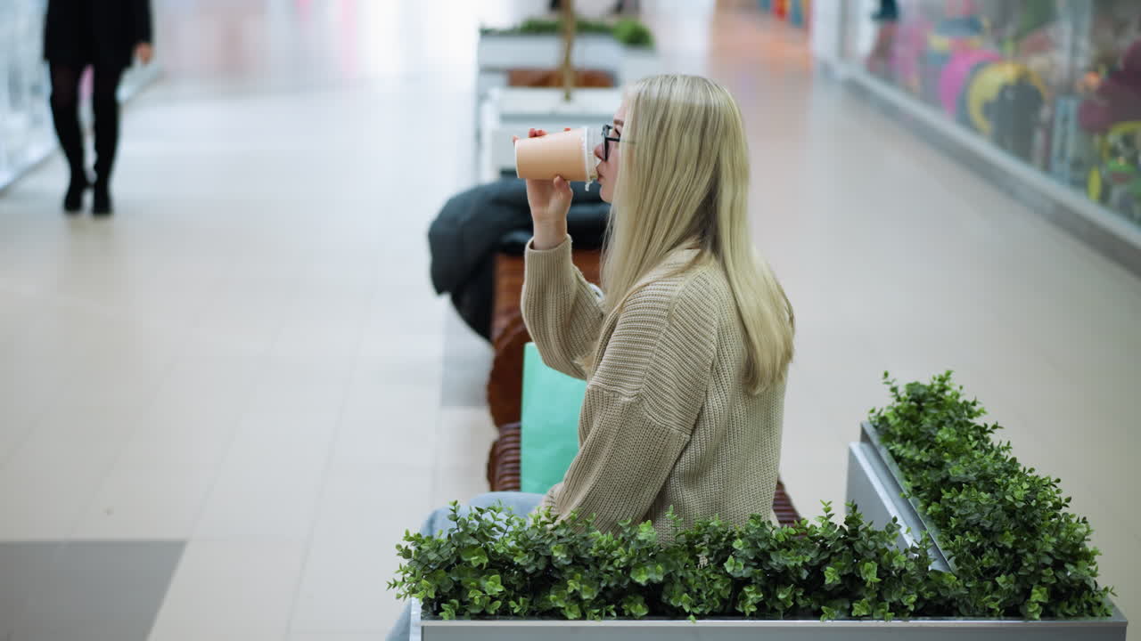 карьерная женщина глотает чай и задумчиво останавливается с декоративными растениями поблизости, размытый фон показывает других покупателей в оживленном торговом центре