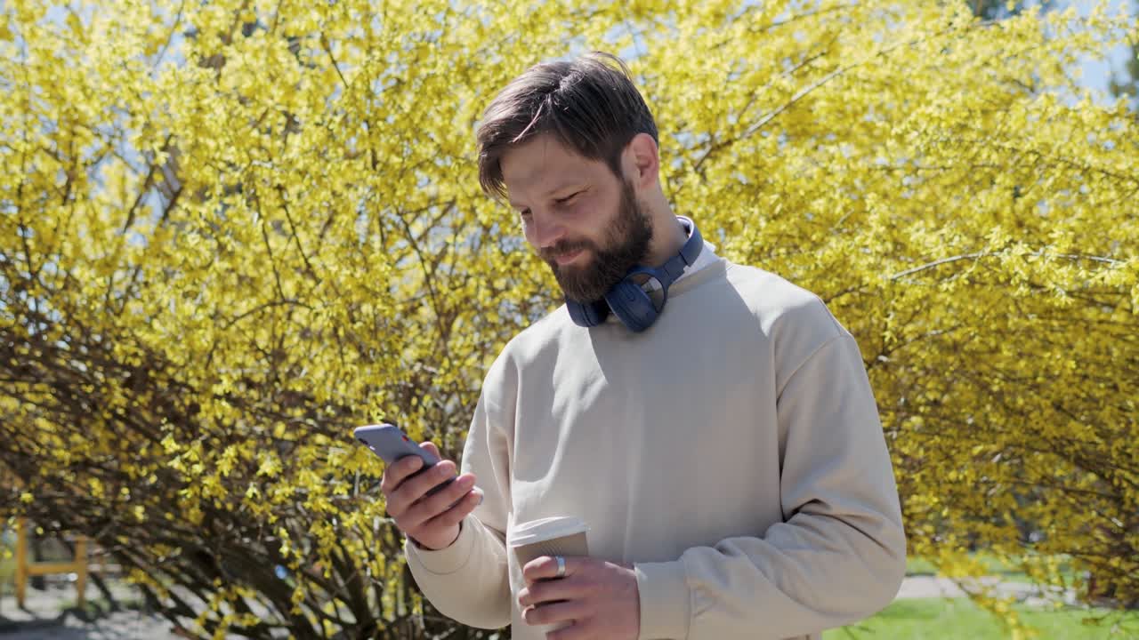 길거리에서 스마트폰을 사용하는 잘생긴 남성, 캐주얼 옷을 입고, 헤드폰을 들고 커피를 들고 휴대 전화로 타이핑하면서 소셜 미디어를 스크롤합니다.