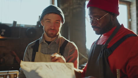 two blacksmiths discussing drawing on paper in sunlit workshop