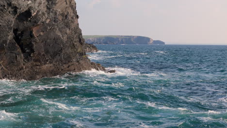 Weathered-sea-cliffs-with-lapping-waves-on-rocky-Cornish-coastline,-aerial