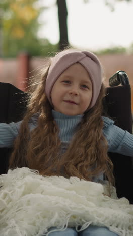 toothless little girl with spinal injury looks at mom holding hand. woman with daughter in wheelchair walks on road in park on sunny day closeup