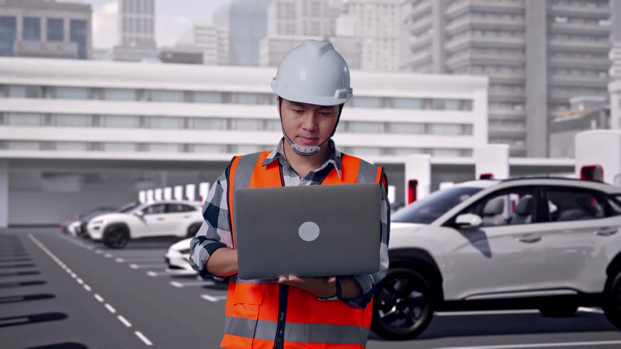 Ingegnere asiatico (uomo) con casco di sicurezza che lavora al laptop in piedi, veicoli elettrici in carica presso una moderna stazione di ricarica in città.