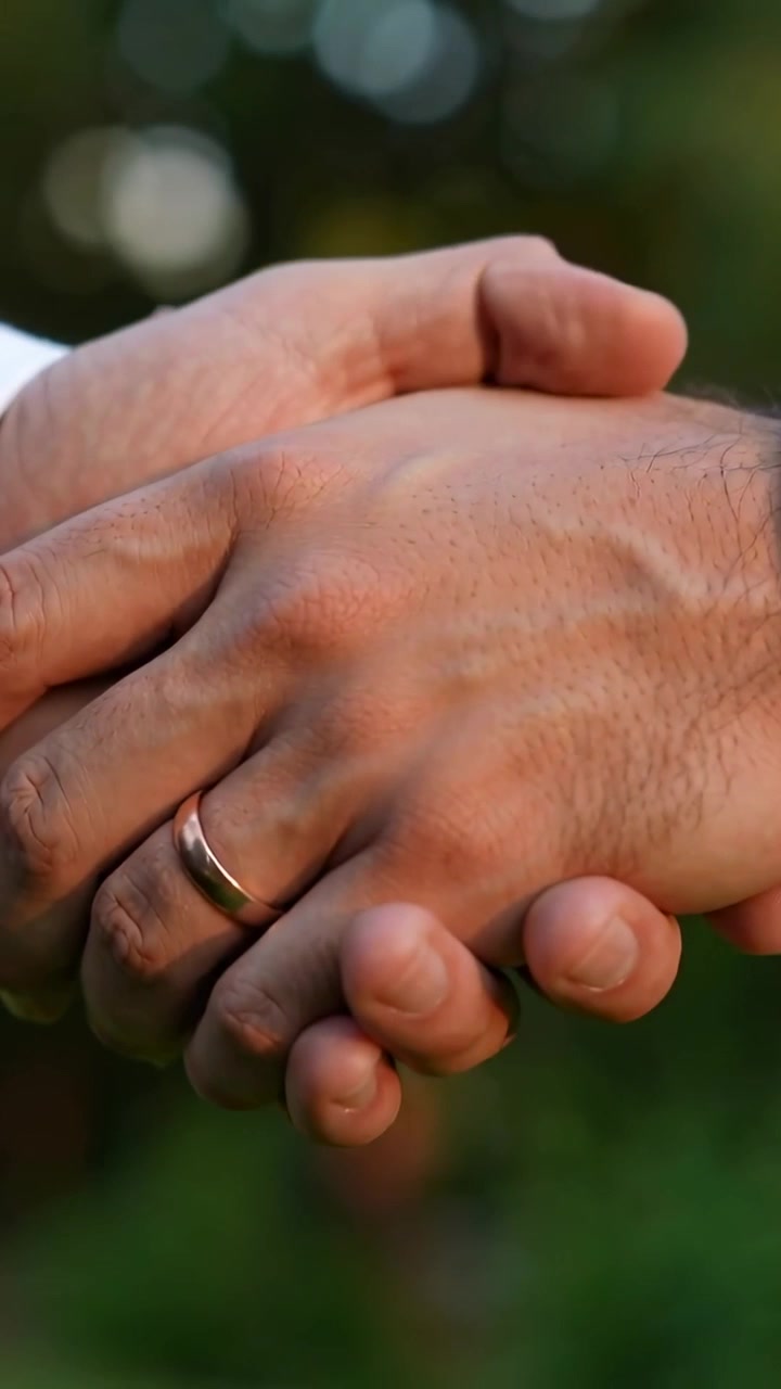 Video verticale: Due uomini si tengono per mano al parco, cercando conforto, con anello di metallo e polsino bianco in evidenza.