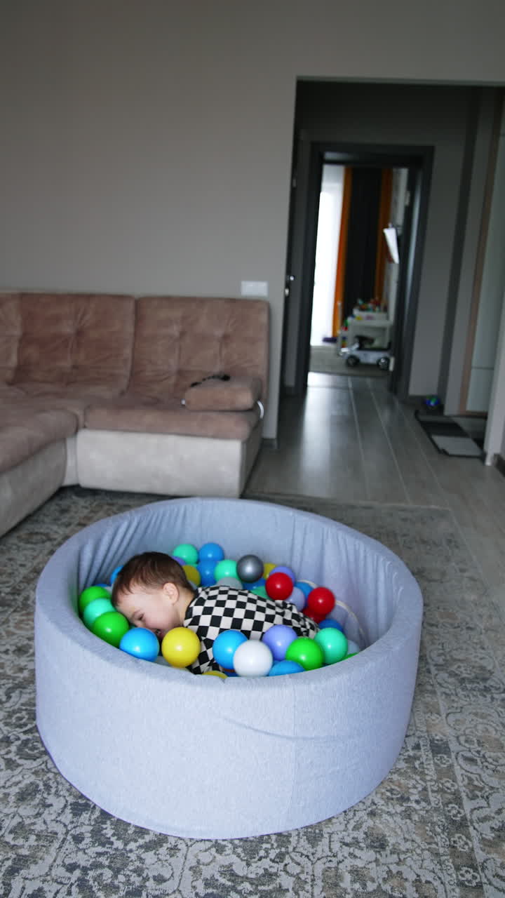 Happy energetic toddler runs up and jumps into the dry pool. Baby boy playing cheerfully at home. Vertical video.