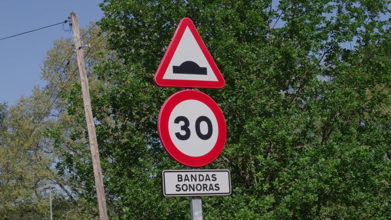 Vista chiara di un dosso stradale e di un segnale stradale di limite 30 km/h etichettato "bandas sonoras", tra alberi verdi, luce solare e cielo blu. Nessuno, inquadratura statica.