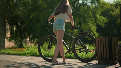 lady mounts bicycle near vibrant garden, adjusting left foot on pedal with focused expression, surrounded by lush greenery, colorful flowers, and tall trees, with distant building view