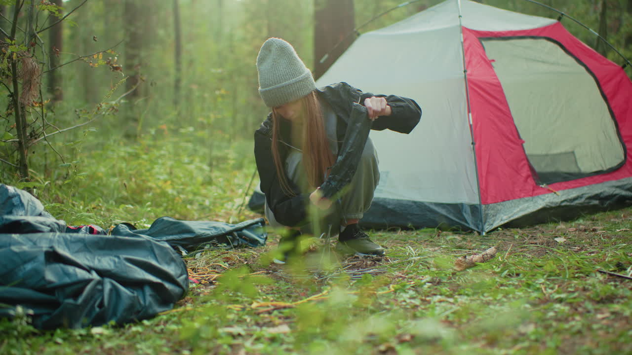 young camper in gray beanie crouches near tent in wooded area picking tent rods from forest floor during quiet outdoor moment surrounded by trees and soft daylight
