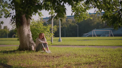 woman sits on grassy ground under tree, hand resting on her head and leg, with sunlight gently reflecting off her, background includes poles and distant stadium