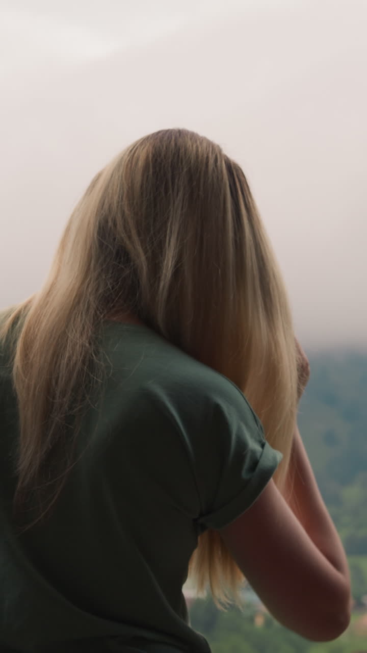 우아한 여성은 긴 금발 머리카락을 고 호텔 객실의 발코니에 서 있으며, 안개와 구름으로 인 오래된 산을 배경으로 느린 동작으로 뒤쪽을 볼 수 있습니다.